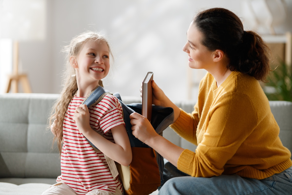 Jeune maman déposant un cahier dans le sac à dos de sa fille souriante.