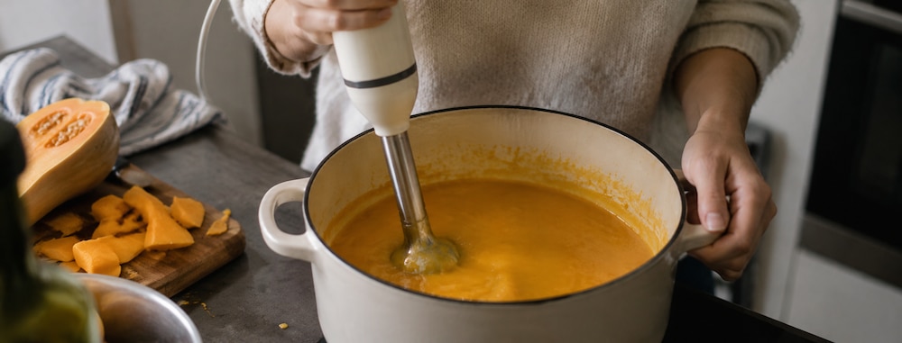Une personne mixe une soupe de butternut dans une cocotte à l'aide d'un mixeur plongeant.