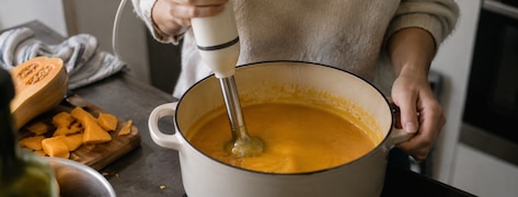 Une personne mixe une soupe de butternut dans une cocotte à l'aide d'un mixeur plongeant.