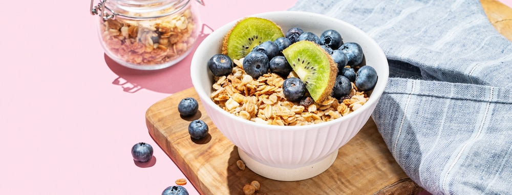  Granola ou muesli au four dans un bol avec kiwi et bleuets sur une planche en bois sur fond rose.