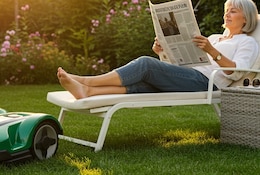 Femme lisant un journal en étant allongée sur un transat dans son jardin pendant qu'un robot tondeuse tond sa pelouse.