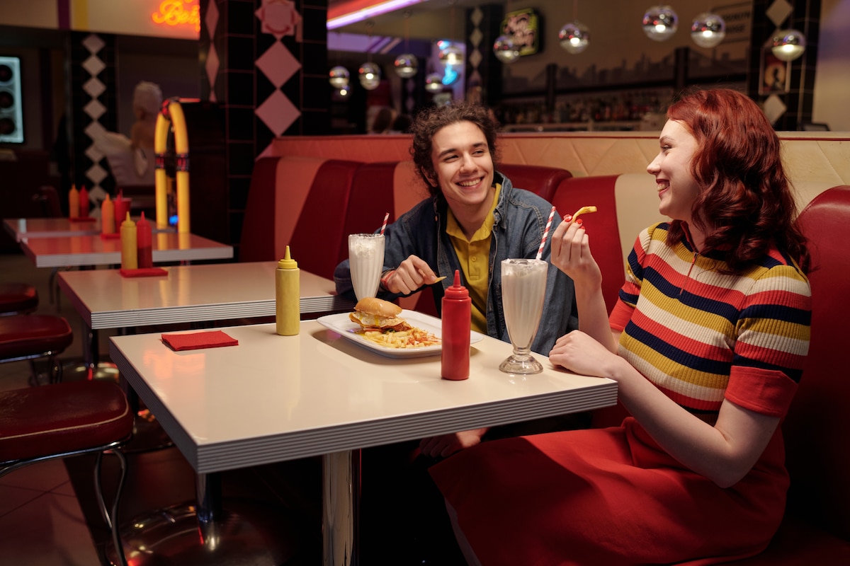 Deux amis dégustent des milkshakes et des burgers dans un diner américain classique avec des banquettes rouges, des éléments décoratifs et un éclairage tamisé.